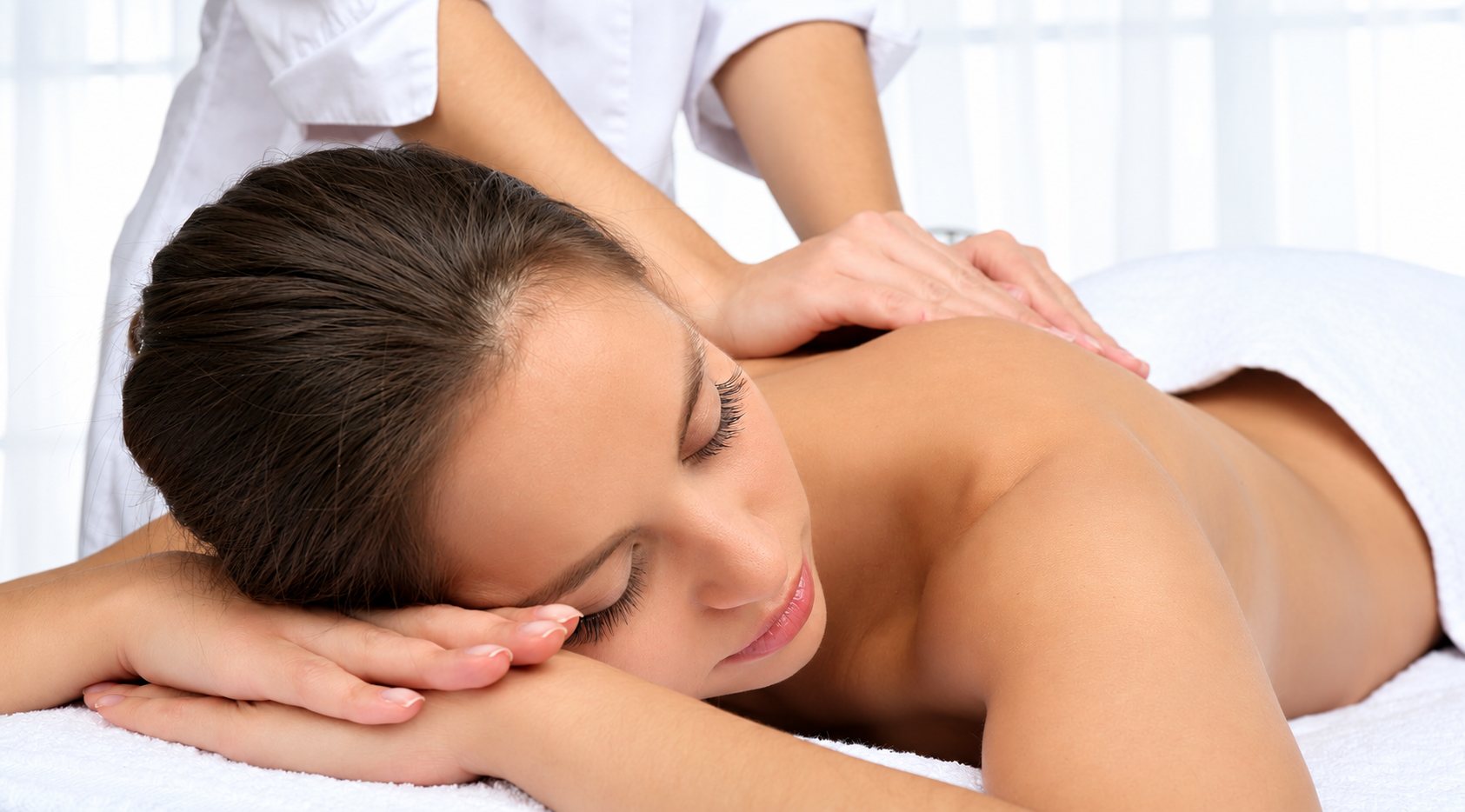 Jeune femme allongée sur une serviette blanche recevant un massage du dos d’une praticienne en blouse blanche.
