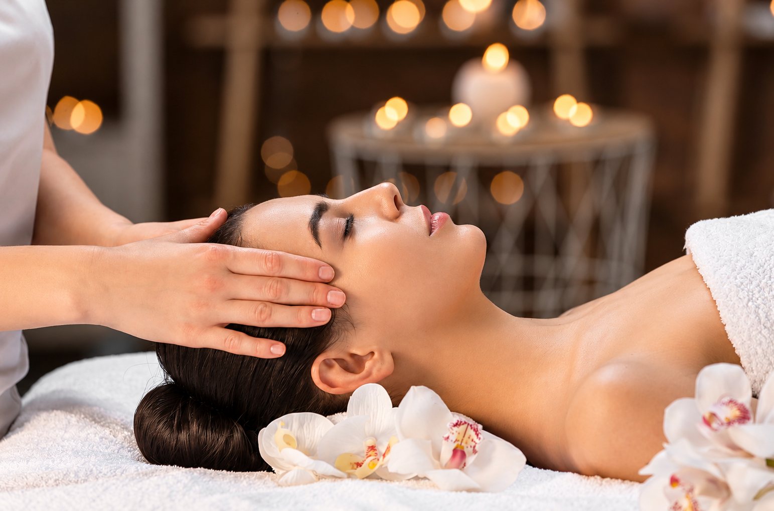 Une femme allongée reçoit un massage du visage, bougies et orchidées blanches en arrière-plan.